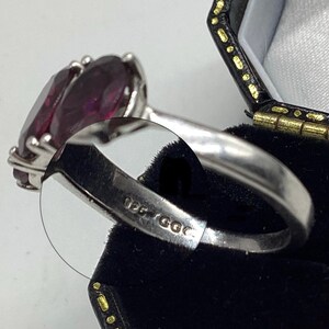 May include: A silver ring featuring a large, oval-cut deep purple gemstone held by four prongs. The inner band of the ring is clearly stamped with the text 925 GGC, indicating the metal composition and maker's mark on the polished silver surface.