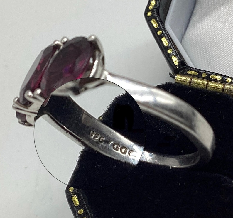 May include: A silver ring featuring a large, oval-cut deep purple gemstone held by four prongs. The inner band of the ring is clearly stamped with the text 925 GGC, indicating the metal composition and maker's mark on the polished silver surface.