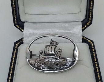 May include: A silver oval brooch featuring a detailed depiction of a ship sailing on the water.