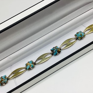以下が含まれることがあります： 金色の金属にターコイズの石がセットされたシルバーのブレスレット。ブレスレットは、黒い裏地が付いた白い箱に展示されています。