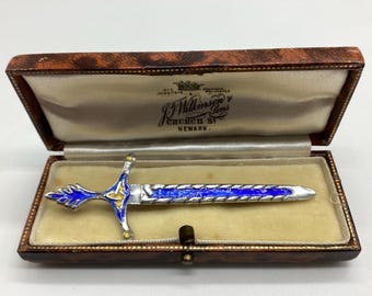 May include: A silver sword brooch with blue enamel and gold accents, displayed in a brown leather box with a white satin lining. The box has a label that reads "J. Wilkinson & Sons, Church St. Newark."