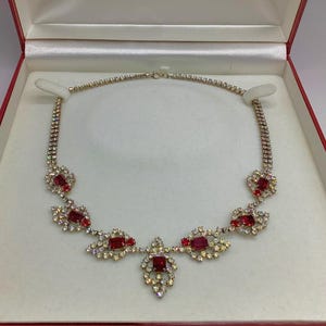 Op de afbeelding: Een sprankelende ketting met rode en heldere edelstenen, gepresenteerd in een rode en witte sieradendoos. De ketting heeft een reeks rode rechthoekige stenen omringd door heldere, ronde stenen.