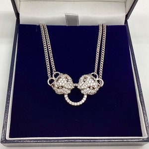 Könnte beinhalten: Eine silberne Halskette mit einer doppelten Kette und einem Anhänger mit zwei Löwenköpfen mit Kristall-Akzenten. Der Anhänger ist durch einen Kreis aus Kristallen verbunden.