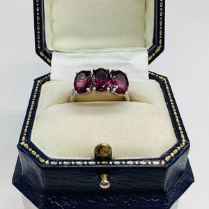 May include: A silver ring featuring three oval-cut deep red gemstones set in a row. The ring is displayed inside a dark blue, octagonal-shaped jewellery box with a white satin interior lining and gold-coloured trim along the edges of the lid.