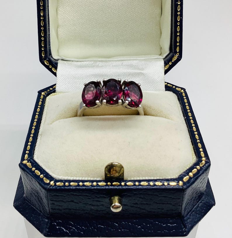 May include: A silver ring featuring three oval-cut deep red gemstones set in a row. The ring is displayed inside a dark blue, octagonal-shaped jewellery box with a white satin interior lining and gold-coloured trim along the edges of the lid.