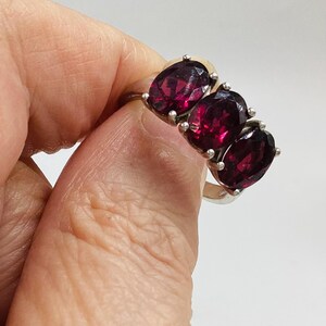 May include: A silver ring featuring three oval-cut, deep red garnet gemstones arranged in a horizontal row. The stones are set in individual silver prongs, creating a continuous cluster design across the top of the polished silver band.