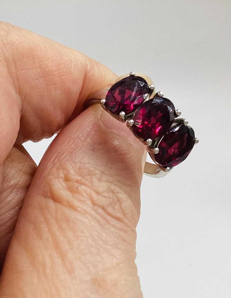 May include: A silver ring featuring three oval-cut, deep red garnet gemstones arranged in a horizontal row. The stones are set in individual silver prongs, creating a continuous cluster design across the top of the polished silver band.