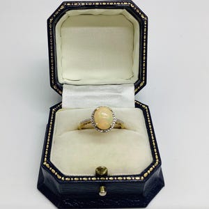 May include: A gold ring featuring an oval opal gemstone, encircled by small clear stones, is presented in a black jewellery box with a cream interior. The ring is the focal point, highlighting its design and details.