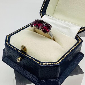 May include: A silver ring featuring three oval-cut deep red gemstones set in a prong setting. The ring is displayed inside a dark blue, octagonal-shaped jewellery box with a cream-coloured interior lining and gold-coloured trim along the edges.