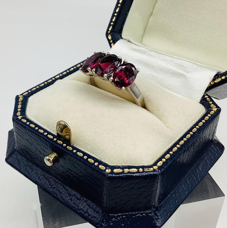 May include: A silver ring featuring three oval-cut deep red gemstones set in a prong setting. The ring is displayed inside a dark blue, octagonal-shaped jewellery box with a cream-coloured interior lining and gold-coloured trim along the edges.