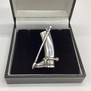 Może przedstawiać: Srebrny wisiorek w kształcie buta jeździeckiego z batem. Wisiorek jest prezentowany w czarnym aksamitnym pudełku.