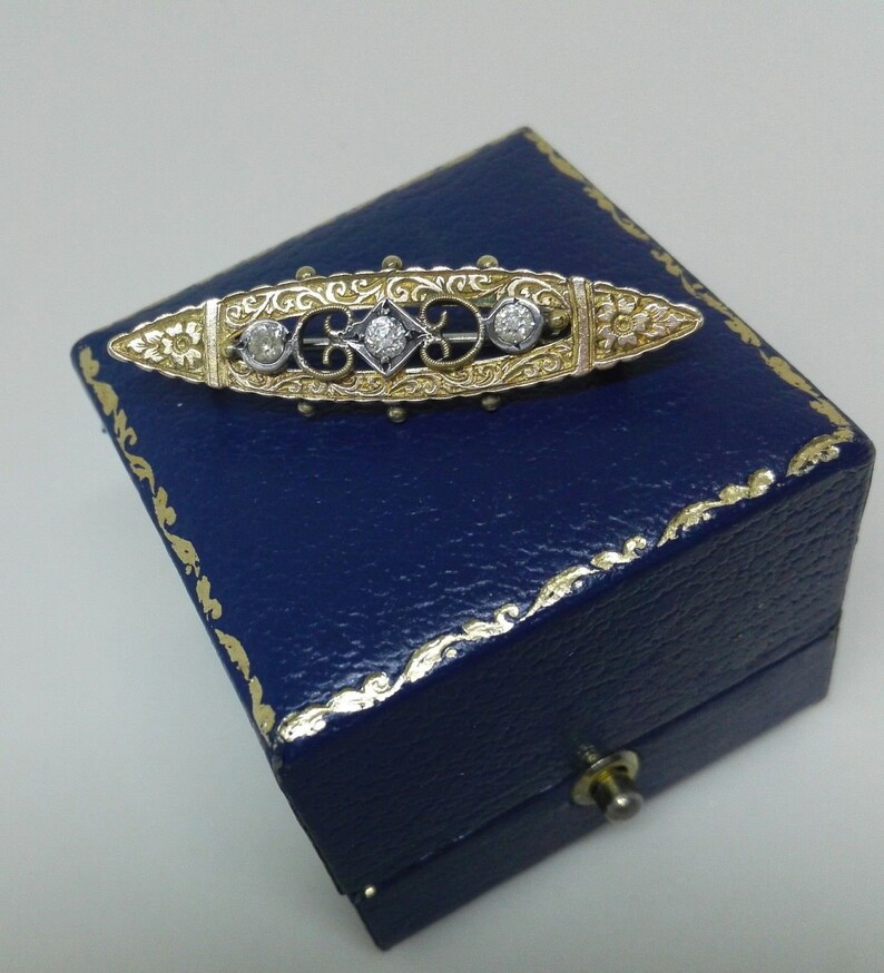 May include: A gold and silver brooch with a delicate floral design and three round diamonds. The brooch is displayed in a blue velvet box.
