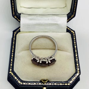 May include: A silver-toned ring featuring three oval-cut dark red gemstones set in a row. The ring is displayed inside a dark blue, octagonal-shaped jewellery box with a cream-coloured velvet interior lining and gold-coloured trim along the edges.