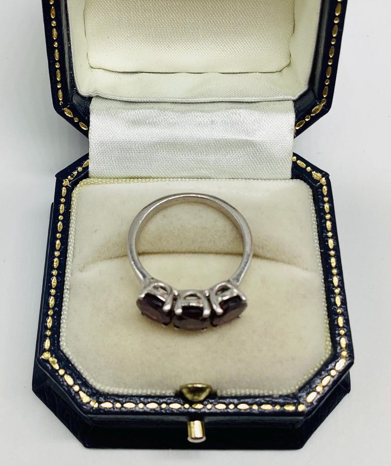 May include: A silver-toned ring featuring three oval-cut dark red gemstones set in a row. The ring is displayed inside a dark blue, octagonal-shaped jewellery box with a cream-coloured velvet interior lining and gold-coloured trim along the edges.