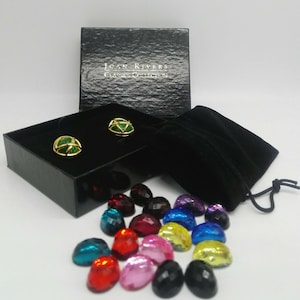 Puede incluir: Un par de pendientes de tono dorado con piedras verdes en una caja negra con una bolsa de terciopelo negro y una variedad de piedras preciosas sueltas en diferentes colores.