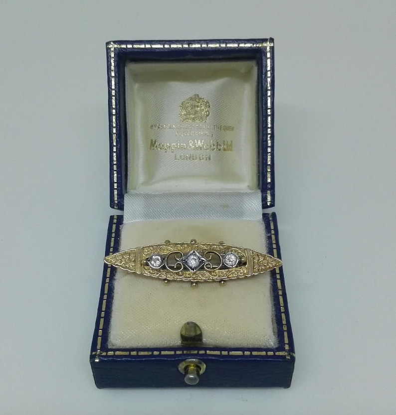 May include: A gold and silver brooch with three diamonds set in a delicate filigree design. The brooch is in a blue velvet box with the inscription "Mappin & Webb London".