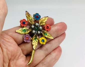 May include: A floral brooch with gold-toned leaves and colorful enamel flowers in red, blue, yellow, and purple. The center features faux pearls and a green gem. The brooch is held in a hand.