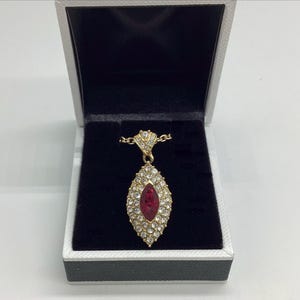 May include: A gold-tone pendant necklace featuring a marquise-shaped centerpiece with a central red stone surrounded by clear pavé crystals. The pendant is suspended from a gold-tone chain and is presented inside a white jewelry box with black lining.