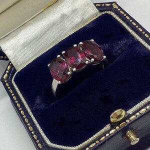 May include: A silver ring featuring three oval-cut, deep red gemstones set in a row. The ring is displayed inside a dark blue velvet-lined jewellery box with a cream-coloured satin interior lining and gold-toned trim along the edges of the box.
