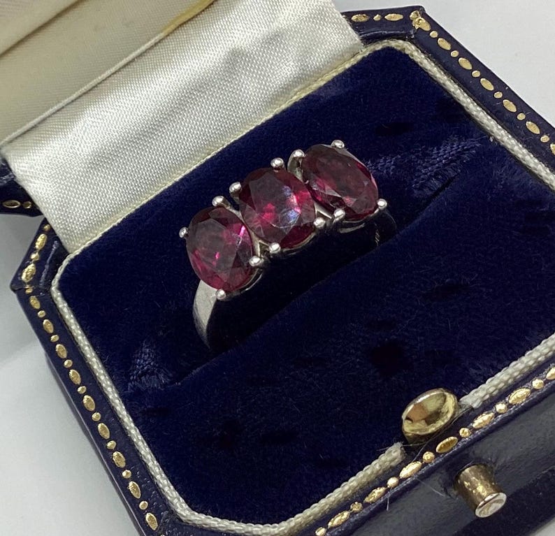 May include: A silver ring featuring three oval-cut, deep red gemstones set in a row. The ring is displayed inside a dark blue velvet-lined jewellery box with a cream-coloured satin interior lining and gold-toned trim along the edges of the box.