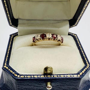 Pode incluir: Um anel de ouro com três pedras preciosas redondas de cor vermelho-escura, alternadas com pequenas pedras claras cravadas numa banda de metal decorativa. O anel é apresentado dentro de uma caixa de joias azul-escura com dobradiças.
