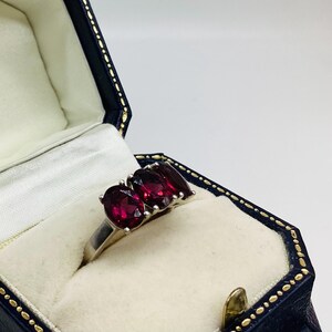May include: A silver ring featuring three oval-cut deep red garnet gemstones set in a row. The ring is displayed inside a dark blue, hexagonal-shaped jewellery box with a cream-coloured interior lining and gold-coloured trim along the edges.