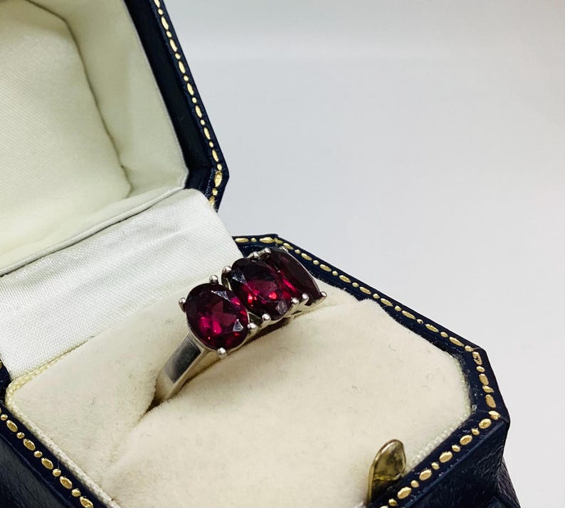 May include: A silver ring featuring three oval-cut deep red garnet gemstones set in a row. The ring is displayed inside a dark blue, hexagonal-shaped jewellery box with a cream-coloured interior lining and gold-coloured trim along the edges.