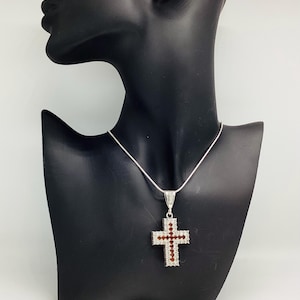 May include: A silver pendant necklace with a cross pendant. The cross is embellished with red and clear gemstones, set in a silver frame. The necklace is displayed on a black mannequin.