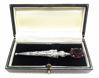 May include: A silver brooch in the shape of a dagger with a large, dark red gemstone at the end. The brooch is in a black velvet lined box.