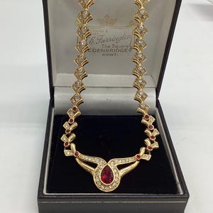 May include: Gold-coloured necklace with a teardrop-shaped red gemstone pendant, surrounded by clear crystals. The necklace features diamond-shaped links with clear crystals and red accents. Presented in a black jewellery box.