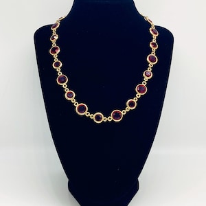 Può includere: Una collana dorata con una serie di grandi pietre preziose rotonde color rosso rubino. La collana è esposta su un manichino nero, evidenziando il design e il contrasto cromatico del gioiello. Le pietre sono distanziate uniformemente e incastonate in una lunetta dorata.