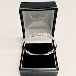 Puede incluir: Una pulsera de plata para bebé con un patrón geométrico, presentada en una caja negra. La pulsera es ajustable y tiene un acabado brillante. El interior de la caja es de terciopelo negro y el forro es plateado.