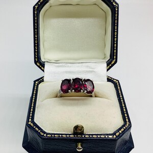 May include: A silver-toned ring featuring three oval-cut, deep red gemstones set in a row. The ring is displayed inside an octagonal, dark blue jewellery box with a cream-coloured velvet interior and gold-coloured trim along the edges.