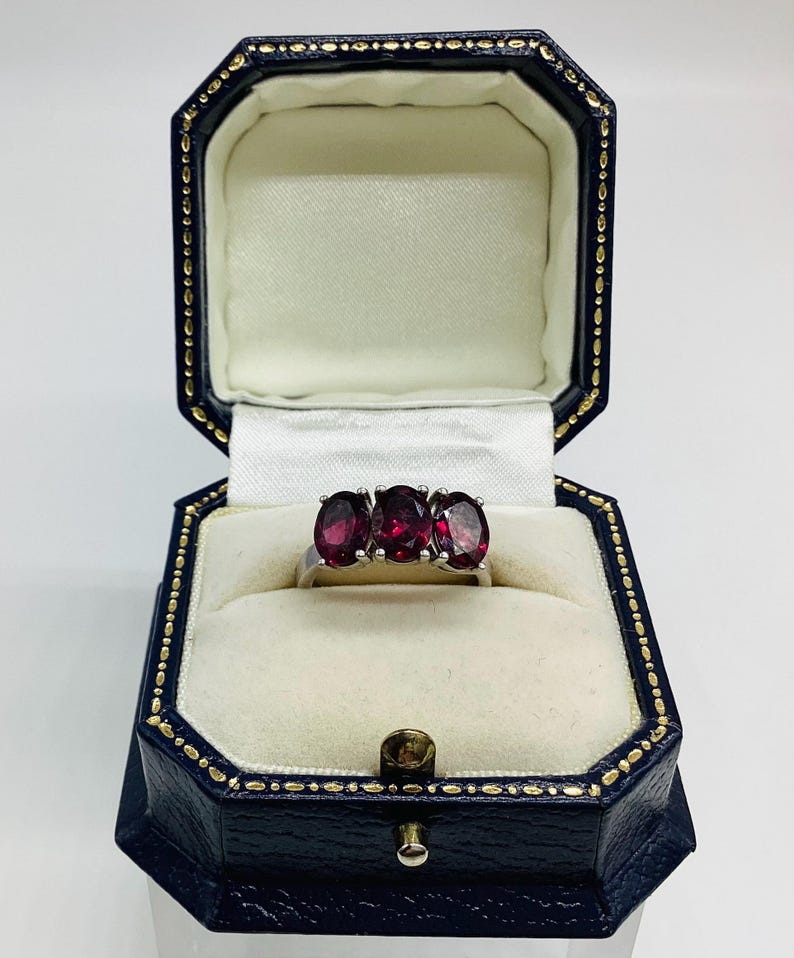 May include: A silver-toned ring featuring three oval-cut, deep red gemstones set in a row. The ring is displayed inside an octagonal, dark blue jewellery box with a cream-coloured velvet interior and gold-coloured trim along the edges.