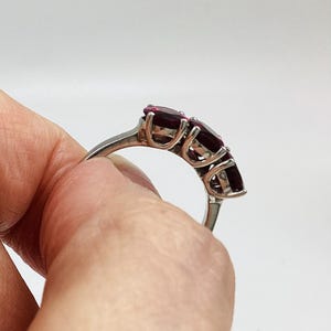 May include: A silver-toned ring featuring three oval-cut deep red gemstones set in a prong setting. The band is smooth and metallic, showcasing the side profile of the three-stone arrangement with visible metal prongs securing each dark red stone.