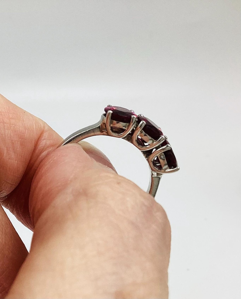 May include: A silver-toned ring featuring three oval-cut deep red gemstones set in a prong setting. The band is smooth and metallic, showcasing the side profile of the three-stone arrangement with visible metal prongs securing each dark red stone.