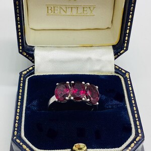May include: A silver-toned ring featuring three oval-cut deep red gemstones set in a row. The ring is displayed inside a dark blue velvet-lined jewellery box with the text & BENTLEY printed in gold on the cream-coloured interior lid lining.
