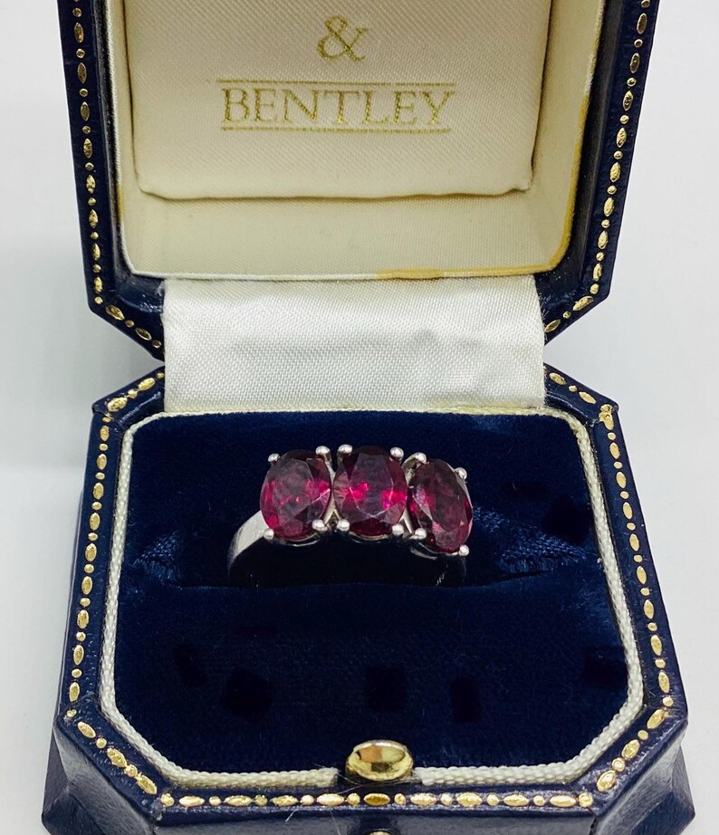 May include: A silver-toned ring featuring three oval-cut deep red gemstones set in a row. The ring is displayed inside a dark blue velvet-lined jewellery box with the text & BENTLEY printed in gold on the cream-coloured interior lid lining.