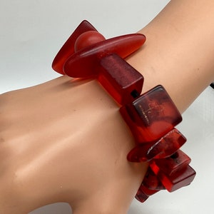 Può includere: Braccialetto in resina rossa con forme geometriche, tra cui una sfera, triangoli e quadrati. Il braccialetto è realizzato da "Arctic" e ha un'etichetta "Made in China".