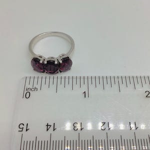 May include: A silver-toned ring featuring three oval-cut deep purple stones set in a row. The ring is positioned above a clear plastic ruler, showing the width of the stone setting is approximately 1.9 centimetres across the top of the band.