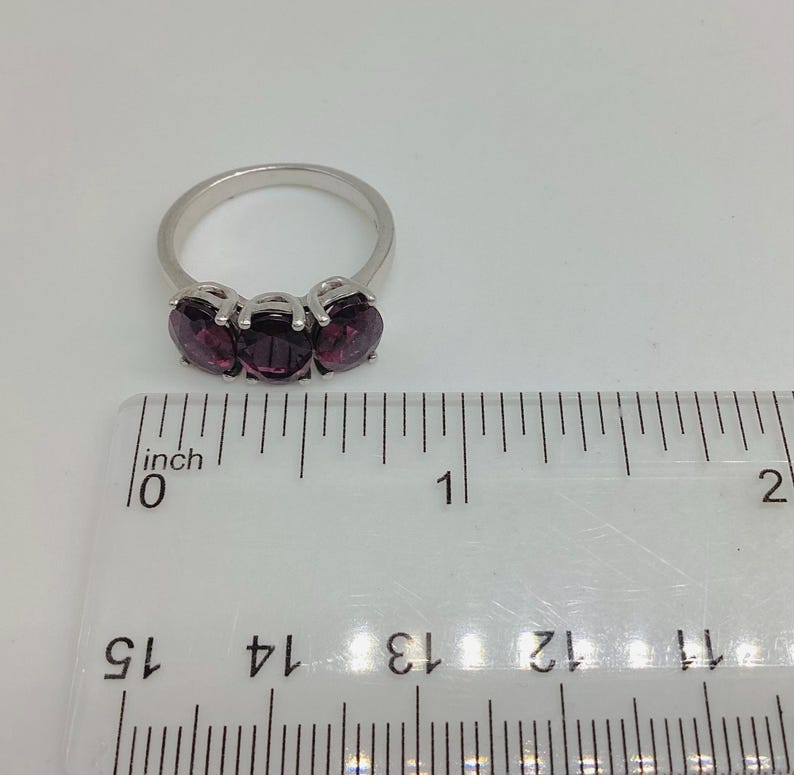 May include: A silver-toned ring featuring three oval-cut deep purple stones set in a row. The ring is positioned above a clear plastic ruler, showing the width of the stone setting is approximately 1.9 centimetres across the top of the band.