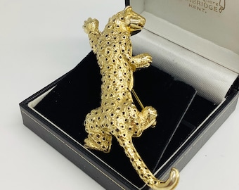 May include: A gold-toned leopard brooch with detailed spots and a textured surface. The brooch is displayed in a black box with a velvet interior, showcasing the jewelry's design and craftsmanship. The brooch is a classic accessory.