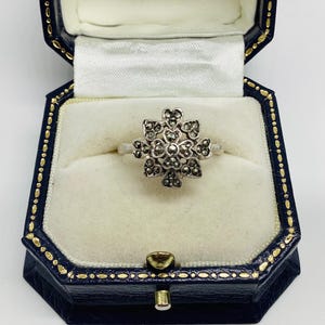 Op de afbeelding: Een zilverkleurige ring met een centraal bloemvormig cluster bezet met talloze kleine, donkere, gefacetteerde stenen. De ring wordt gepresenteerd in een donkerblauw, achthoekig juwelendoosje met een witte, gevoerde binnenkant.