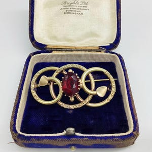 Op de afbeelding: Een gouden broche met drie in elkaar grijpende cirkelvormige ringen met gegraveerde patronen en een centrale ovaal geslepen dieprode edelsteen. De broche wordt gepresenteerd in een vintage sieradendoosje met een donkerblauwe fluwelen voering.