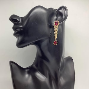 Può includere: Orecchini pendenti dorati con dettagli di pietre preziose rosse. Ogni orecchino presenta una gemma rossa a forma di goccia nella parte inferiore e una gemma rossa a forma di cuore nella parte superiore. Il design include elementi a forma di freccia con piccole gemme trasparenti.