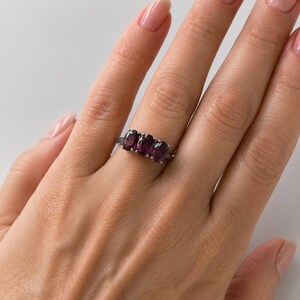 May include: A silver-toned ring featuring three oval-cut, deep purple gemstones set horizontally in a row. The band is thin and metallic, showcasing a polished finish that complements the dark, faceted stones arranged across the centre.