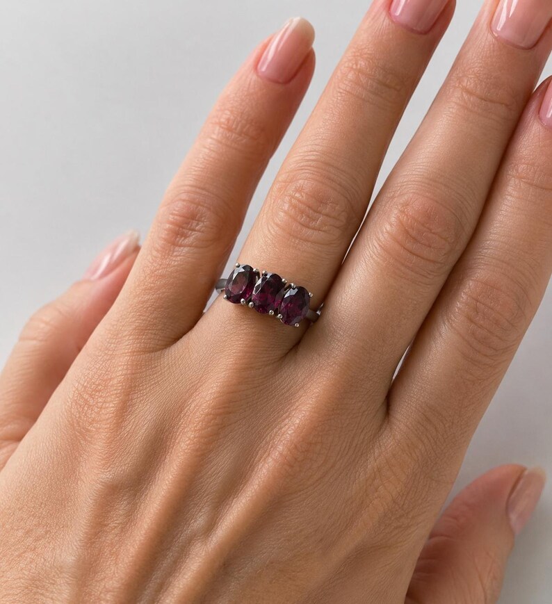 May include: A silver-toned ring featuring three oval-cut, deep purple gemstones set horizontally in a row. The band is thin and metallic, showcasing a polished finish that complements the dark, faceted stones arranged across the centre.