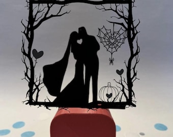 Può includere: Un cake topper nuziale a silhouette nera raffigurante una coppia che si bacia all'interno di una cornice di rami d'albero. Sono inclusi anche una ragnatela, cuori e una zucca. Il topper è montato su un blocco rosso.