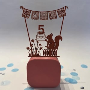 Puede incluir: Adorno de pastel de madera marrón con una ardilla sosteniendo un pastel con el número 5. El adorno incluye una pancarta con recortes decorativos y detalles florales, perfecto para una celebración de quinto cumpleaños.