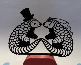 May include: A black laser-cut wedding cake topper featuring two pangolins facing each other. One pangolin wears a top hat, the other a veil. The topper is mounted on a red square base. Decorative confetti is scattered around the base.